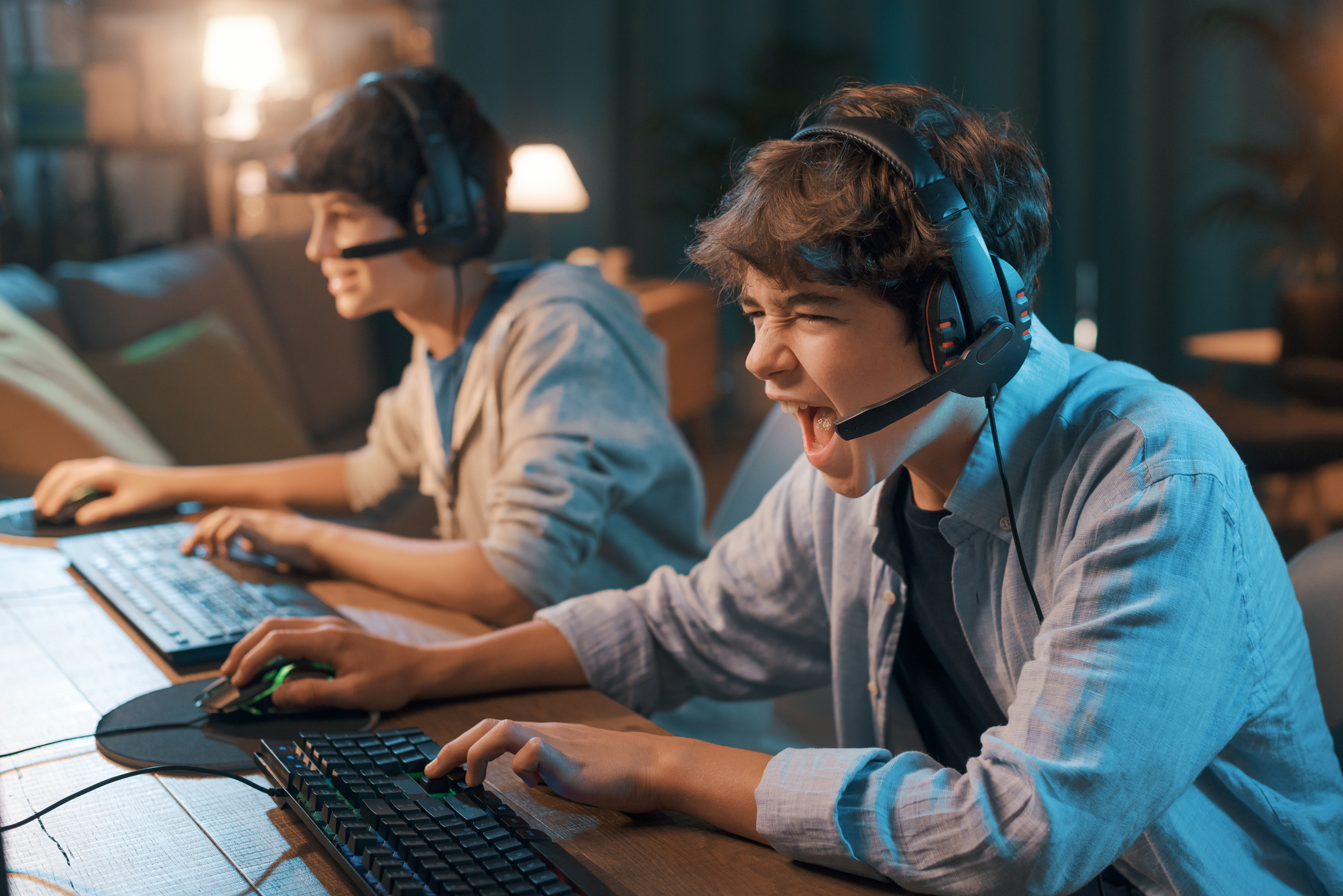 Two young people sitting next to each other playing video games on their computers while wearing headsets.