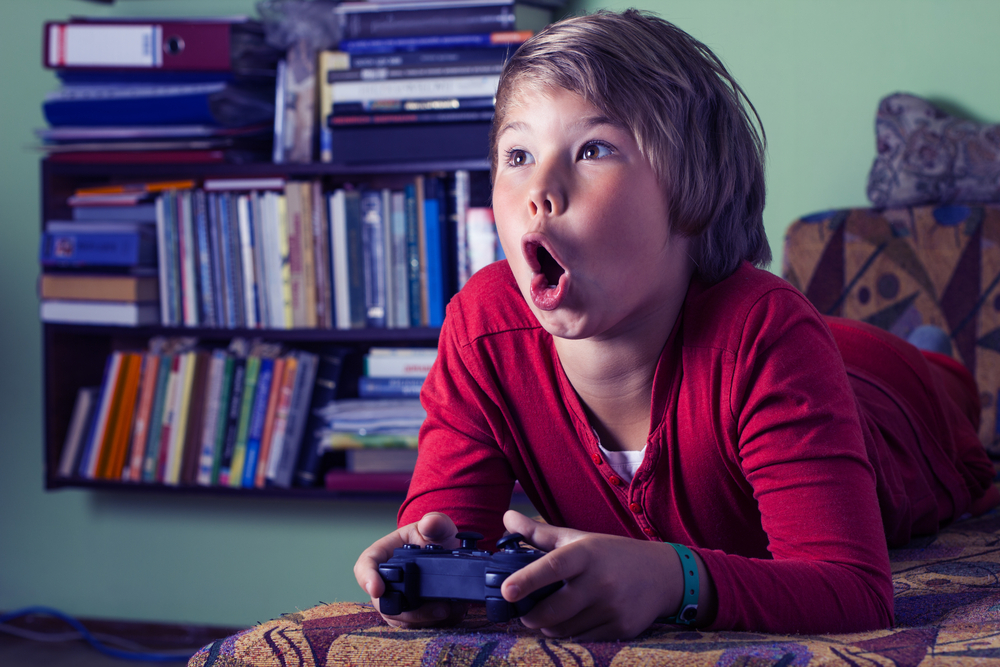 Child laying on bed plays video games with expression of awe.